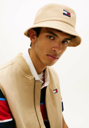 Junger Mann trägt einen beigen Anglerhut und eine Jacke mit Tommy Jeans Logos, schaut direkt in die Kamera vor einem schlichten weißen Hintergrund.