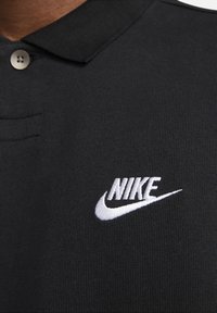 Svart pikétröja med vitt broderat Nike-logotyp och swoosh på bröstet, med krage och knappdetalj.