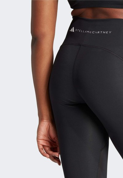 Close-up van een persoon in zwarte high-waisted sportlegging met "Stella McCartney" branding bij de tailleband.