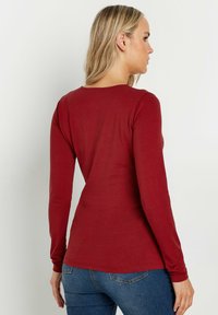 Maglia a maniche lunghe, aderente, in tessuto rosso scuro con scollatura rotonda. Texture liscia e cuciture minime, abbinata a jeans blu.