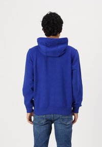 Haut en coton bleu avec une coupe décontractée, poche kangourou et capuche doublée. Les poignets et l'ourlet sont côtelés pour un ajustement confortable. Porté avec un jean en denim.