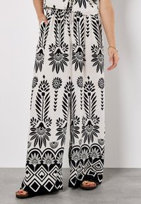 Pantalons larges noir et blanc avec des motifs floraux et géométriques, dotés d'une taille élastique et de poches. Le tissu semble léger.