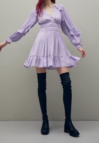 Lavendelkleurige jurk met gerimpelde mouwen, een getailleerde taille en een uitlopende rok. Gecombineerd met zwarte leren laarzen tot aan de knie en vierkante hakken.