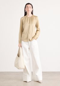 Blusa di seta beige con arricciatura sul colletto, abbottonatura frontale e maniche lunghe; abbinata a pantaloni bianchi a gamba larga e una borsa beige chiaro.