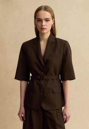 Vrouw met steil blond haar draagt een donkerbruine blazer met riem, drie knopen en korte mouwen tegen een beige achtergrond.