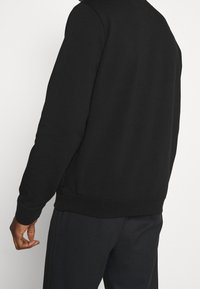 Sudadera negra con cuello redondo, ribete en el dobladillo y los puños, y una textura suave, mostrada desde la parte posterior con una mano descansando al lado.