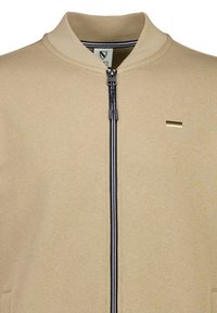 Giacca beige con zip, colletto alto, cerniera a contrasto blu navy e bianca, e un piccolo logo orizzontale sul petto.