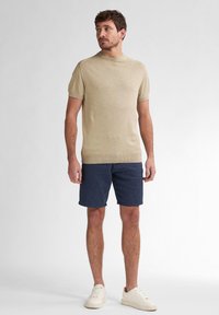 Helle beige Kurzarm-Strickjacke mit Rundhalsausschnitt, kombiniert mit marineblauen Shorts mit Streifen. Das Modell trägt weiße Sneakers. Einfacher und lässiger Look.