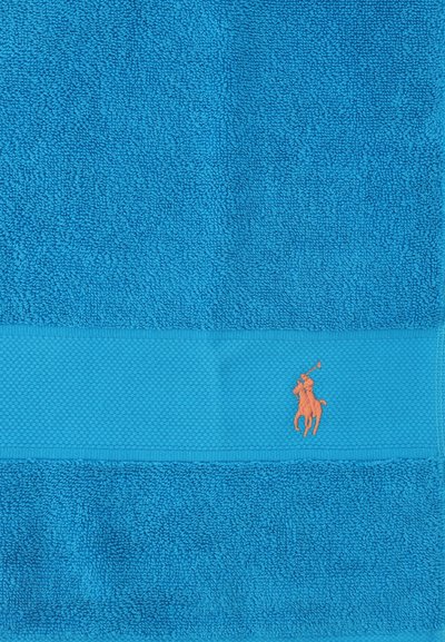 Serviette en éponge bleue avec bande texturée et petit logo brodé représentant un joueur de polo orange près du bord.