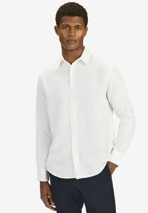 Man som bär en vit långärmad skjorta med knappar, med en hand i fickan och ett neutralt ansiktsuttryck, stående mot en enkel bakgrund.