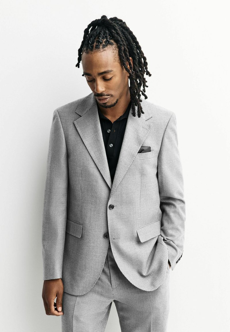 Gray checkered suit with a tailored fit, notched lapels, two front pockets, and a pocket square. Black polo shirt underneath.