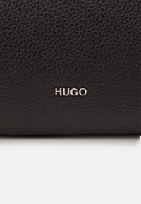 Svart läderplånbok med texturerad yta, som har en silverfärgad "HUGO"-logotyp på framsidan. Enkel design med subtila detaljer.