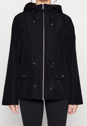 Vrouw draagt een zwarte jas met capuchon, voorzien van een ritssluiting aan de voorkant, verstelbare trekkoorden bij de taille en capuchon, en twee klepzakken met knopen aan de voorkant.