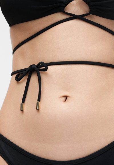 Κοντινό πλάνο στη μέση, με μαύρο μπικίνι με λεπτές τιράντες δεμένες σε φιόγκο και χρυσά άκρα γύρω από τη μέση.