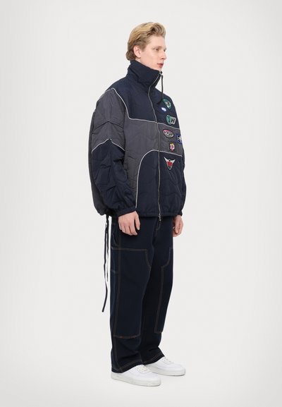 Padded jacket in dark gray and navy with white piping, featuring multiple patches. Paired with navy pants and white sneakers.