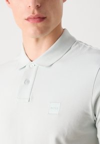 Světle šedé polo tričko s texturovanou tkaninou, dvěma knoflíky a malou bílou nášivkou na levé části hrudi.