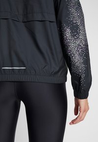 Chaqueta atlética negra con un patrón de puntos blancos y morados en la manga, usada con leggings negros brillantes, mostrando la espalda y la mano derecha.