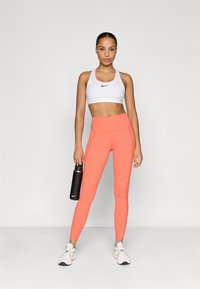 Sujetador deportivo blanco con logo negro, leggings naranjas con costuras laterales y botella de agua negra. Zapatillas ligeras con cordones y suelas texturizadas.