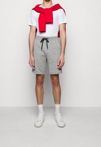 Shorts en coton gris avec un cordon de serrage foncé, associés à un t-shirt blanc et un pull rouge porté négligemment autour des épaules, le tout complété par des baskets blanches.