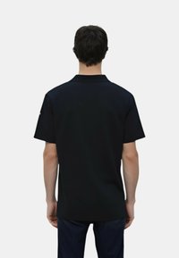 Polo shirt nera a maniche corte, colletto piatto e texture liscia; presenta un logo discreto sulla manica sinistra. Indossata con jeans scuri.