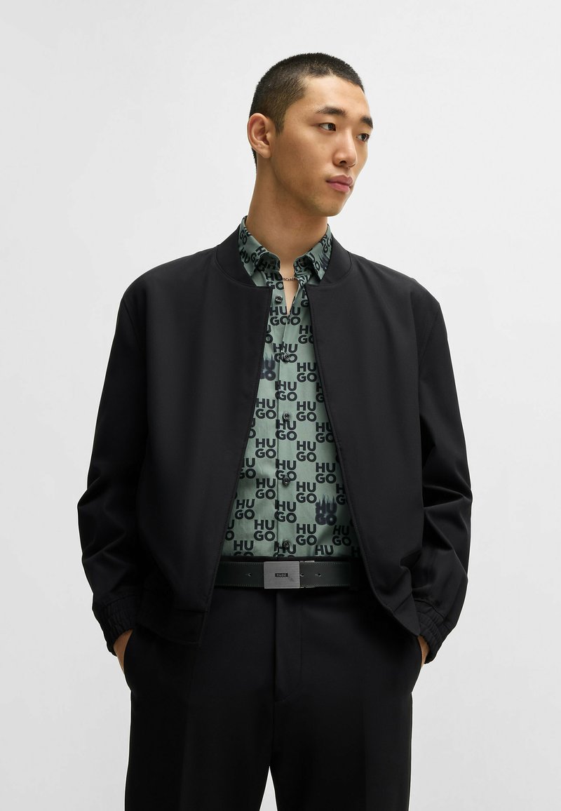 Veste bomber noire avec un col côtelé portée sur une chemise verte à motifs avec un texte sombre. Ceinture noire avec une boucle en argent, pantalons ajustés.