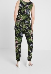 Mono negro con patrón de hojas tropicales en verde y rosa, parte superior sin mangas, cintura elástica y piernas cónicas. Combinado con sandalias de tacón blancas.