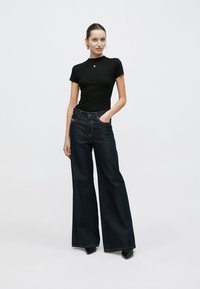 Top nero aderente a maniche corte e jeans in denim scuro a vita alta con gamba ampia e cuciture a contrasto. Il modello indossa tacchi neri a punta.