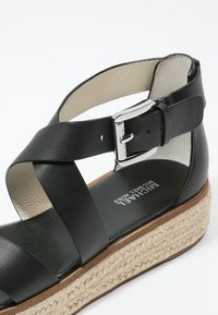 Sandale compensée en cuir noir avec brides croisées, boucle argentée et semelle plateforme en jute tressée, signée Michael Kors sur la semelle intérieure.