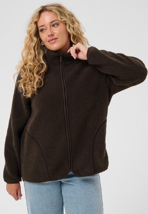 Frau mit lockigem blondem Haar trägt eine dunkelbraune Fleecjacke mit Reißverschluss und hellblaue Jeans und berührt mit der rechten Hand den Kragen.