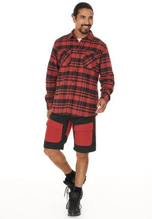 Chemise en flanelle à carreaux rouge et noir avec deux poches poitrine, associée à un short noir hauteur genoux avec des poches cargo rouges et des bottes noires.