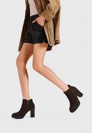 Persona che indossa stivaletti neri con tacco alto, pantaloncini in pelle neri, giacca beige, con una mano in tasca, che cammina su uno sfondo semplice.
