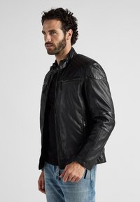 Schwarze Lederjacke mit Reißverschluss, Schulterpolstern und strukturierten Details an den Schultern. Getragen über einem schwarzen Hemd und hellblauen Jeans.