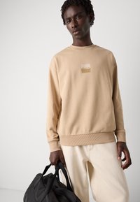 Beige sweatshirt with a square logo in the center, ribbed cuffs and hem, paired with off-white pants. The model holds a black bag.