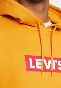 Žlutá mikina s kapucí, na které je červené logo "LEVI'S", s šňůrkou na stažení kapuce a měkkou texturou. Detailní pohled na límec a textilní detaily.