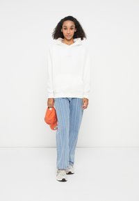 Weißer Kapuzenpullover mit einer Vordertasche, kombiniert mit hellblauen gestreiften Jeans. Das Outfit umfasst eine orangefarbene Mütze und neutrale Sneakers.