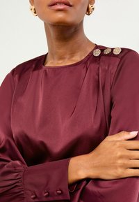 Burgundské saténové blouse s dlhými rukávmi, jemnými záhybmi a tromi ozdobnými gombíkmi na pleci. Priliehavé manžety s gombíkovými detailmi.