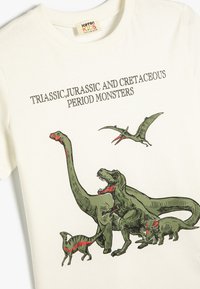 Biała bawełniana koszulka z grafika zielonego dinozaura, w tym T. rexa i inne dinozaury, z czerwonymi akcentami i wyraźnym tekstem powyżej.