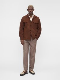 Homme aux cheveux blonds courts portant une veste marron boutonnée, une chemise beige clair, un pantalon taupe et des mocassins bicolores, debout avec une main dans la poche.