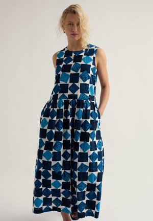 Žena s blond vlasmi oblečená v rukávmi mididievčenských šatách s modrým geometrickým štvorcovým vzorom, stojaca s jednou rukou v vrecku.