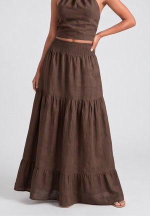 Kobieta ubrana w brązowy bezrękawnik i pasującą warstwową długą spódnicę, stoi z jedną ręką na biodrze.