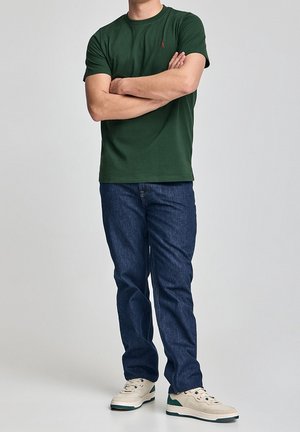 Person wearing a dark green short-sleeve t-shirt, blue jeans, and beige sneakers with green accents, standing with arms crossed.