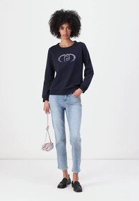 Felpa blu marina con una grafica circolare e a motivi in colori chiari, abbinata a jeans chiari strappati e mocassini neri, mentre tiene una piccola borsa rosa.