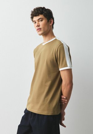 Ung mand stående med hænderne bag ryggen iført brun t-shirt med hvid skulderstribe og sorte bukser mod ensfarvet baggrund.