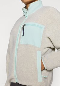 Ζακέτα από φλις σε ανοιχτό γκρι με λαιμόκοψη σε μέντα πράσινη και τσέπη στο στήθος. Διαθέτει φερμουάρ και πλαϊνές τσέπες. Μαλακή υφή.