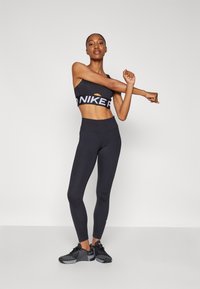 Fekete sportmelltartó, kivágással és logóval, magas derekú fekete leggings, valamint fekete sportcipők texturált talppal, sima háttér előtt.