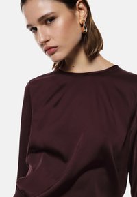 Blusa di seta borgogna con maniche lunghe, scollatura rotonda e superficie liscia e lucida, caratterizzata da sottili dettagli drappeggiati.