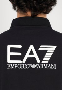 Čierne polo tričko s bielou vyšívanou logom, na ktorom je na chrbte výrazne zobrazený text "EA7 Emporio Armani".