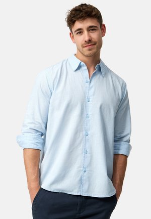 Junger Mann mit kurzen lockigen braunen Haaren, der ein hellblaues Hemd mit hochgekrempelten Ärmeln und dunkle Hose trägt, Hände in den Taschen.