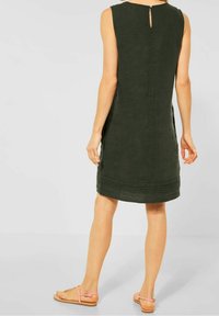 Ärmelloses dunkelgrünes Leinenkleid mit rundem Ausschnitt, Schlüsselhole-Detail am Rücken und dezentem Faltenwurf am Saum. Getragen mit rosa Sandalen mit Riemchen.