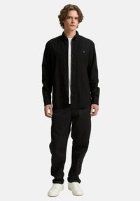 Camisa negra de botones sobre una camiseta blanca, combinada con pantalones negros y zapatillas blancas. Presenta un corte slim, casual y una tela texturizada.